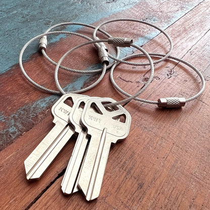 Three keys labeled "KW1" and "USA" are attached to two Wire Loop Luggage Tag Holders - 6 inch Metal Stainless Steel Cable Ring Connectors for Keys or Luggage Tags on a wooden surface, making for multi-functional and secure key organization.