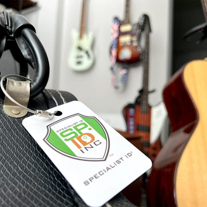 Close-up of a Specialist ID badge attached to a textured bag with various guitars hanging on the wall in the background, secured by Wire Loop Luggage Tag Holders - 6 inch Metal Stainless Steel Cable Ring Connectors for Keys or Luggage Tags.