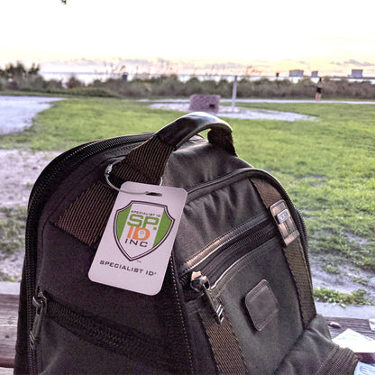 A black backpack with a Wire Loop Luggage Tag Holders - 6 inch Metal Stainless Steel Cable Ring Connectors for Keys or Luggage Tags attached is placed on a wooden bench overlooking a grassy area and a body of water in the background during sunset.